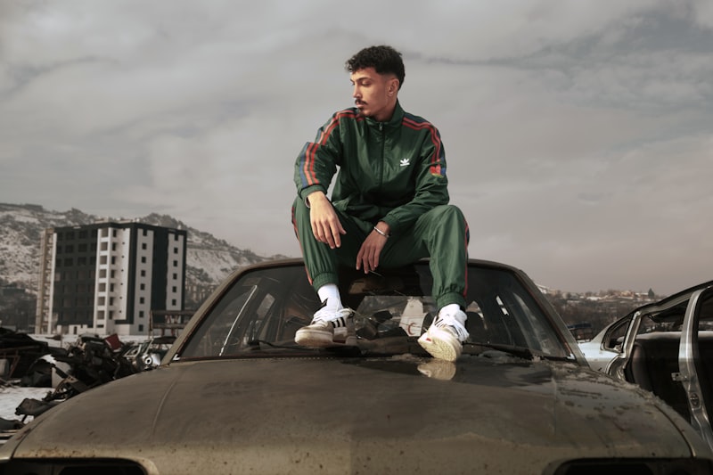 Man in tracksuit sits on car hood, urban decay background.
